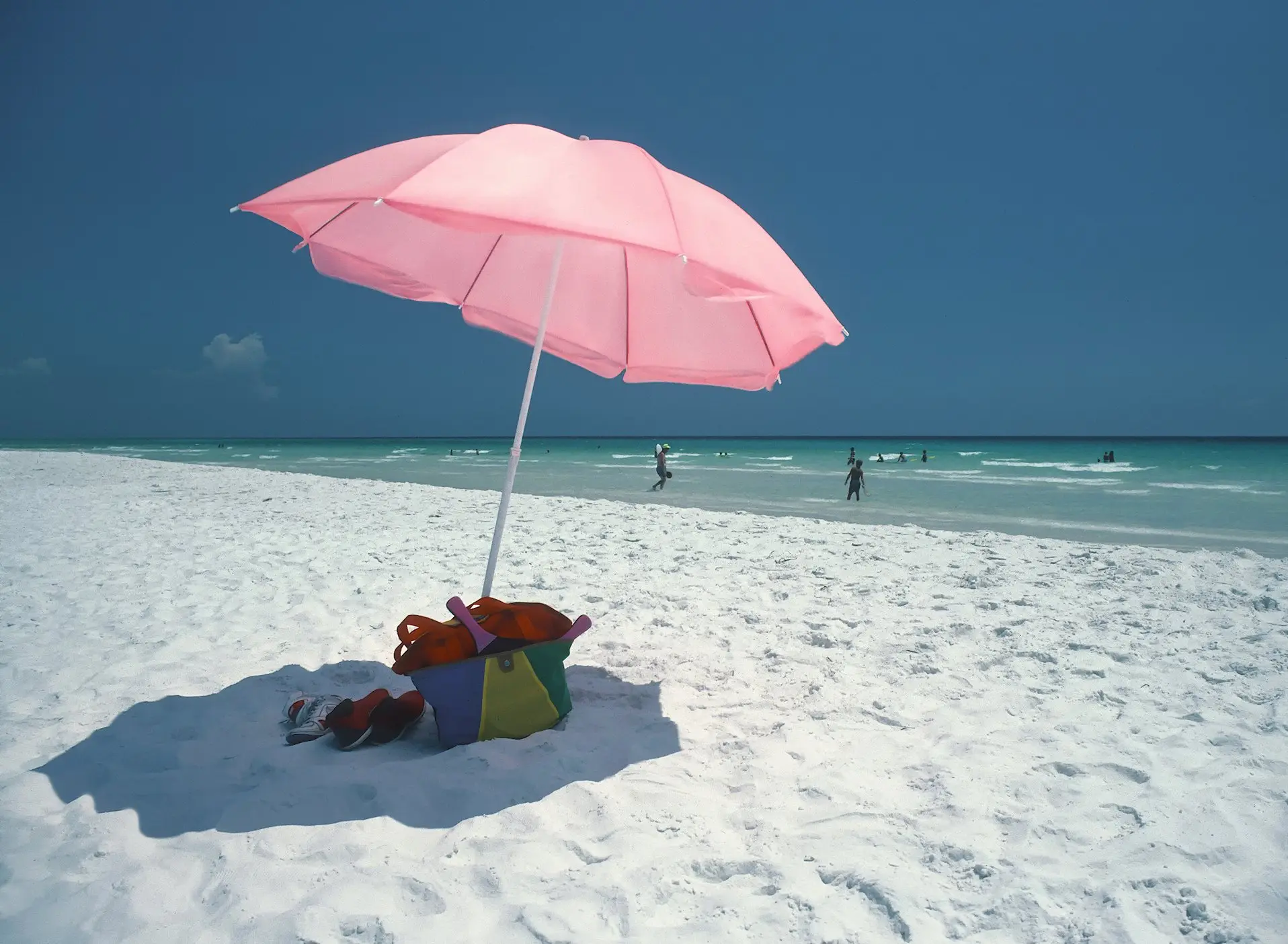 A pink beach umbrella on white sand at Seaside with clear Gulf water behind it